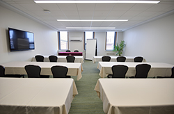 A view of a MSU Union meeting space that has rows of tables with white tablecloths and black chairs, and windows to the front with a television to the left.